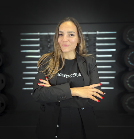 Mujer sonriente con los brazos cruzados, lleva una chaqueta negra, fondo con equipamiento de gimnasio.
